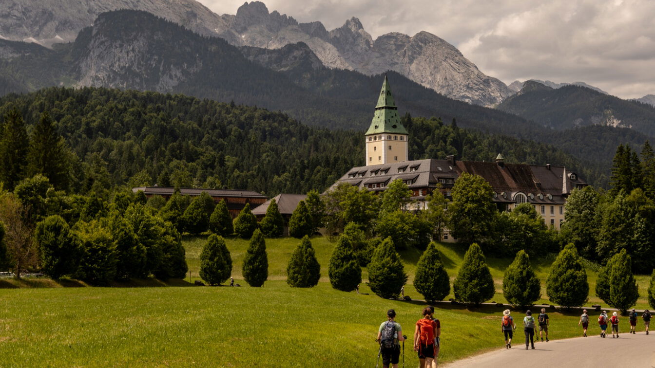 (c) Sonja Koller | 1000things Wanderer auf einem Weg zum Schloss Elmau bei Garmisch-Partenkirchen, umgeben von grüner Natur und den majestätischen Alpen.