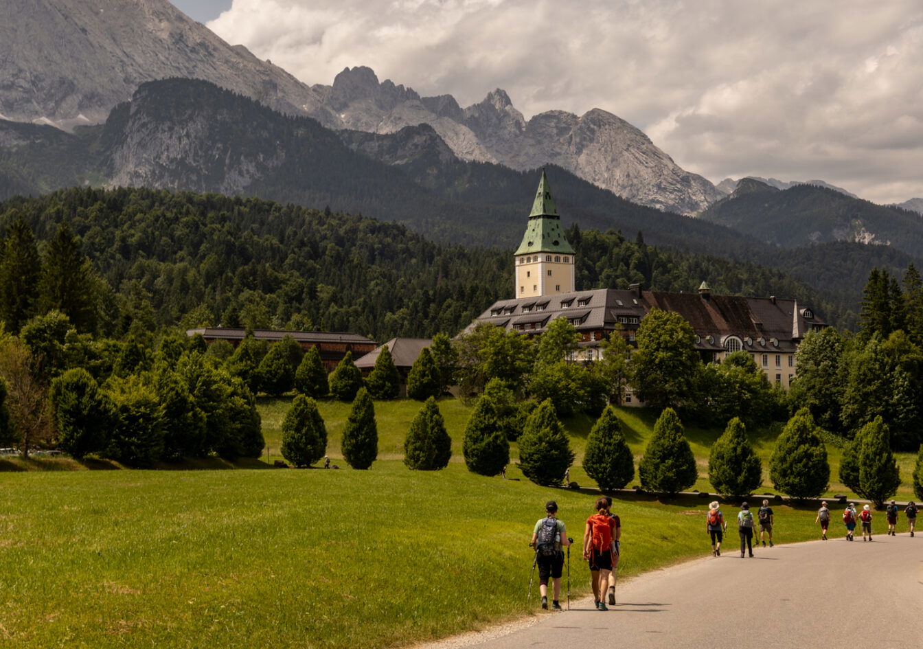 Wanderer auf einem Weg zum Schloss Elmau bei Garmisch-Partenkirchen, umgeben von grüner Natur und den majestätischen Alpen.