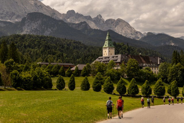 Wanderer auf einem Weg zum Schloss Elmau bei Garmisch-Partenkirchen, umgeben von grüner Natur und den majestätischen Alpen.