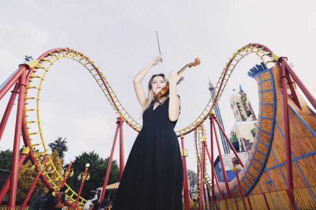 Eine Frau in einem langen schwarzen Kleid spielt Geige im Wiener Prater vor einer markanten rot-gelben Looping-Achterbahn. Im Hintergrund sind weitere Fahrgeschäfte und ein Turm zu sehen. Um sie herum stehen Besucher. Die Szene stammt vom ISA Festival in Wien, das kostenlose Konzerte in ganz Wien bietet.