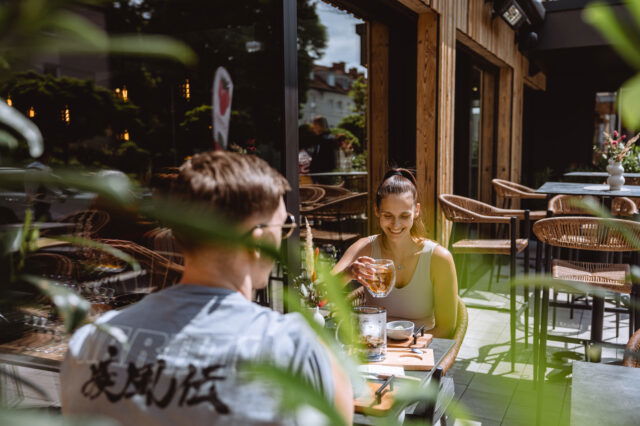 Zwei Menschen auf einer Terrasse, die begrünt und sonnig ist.