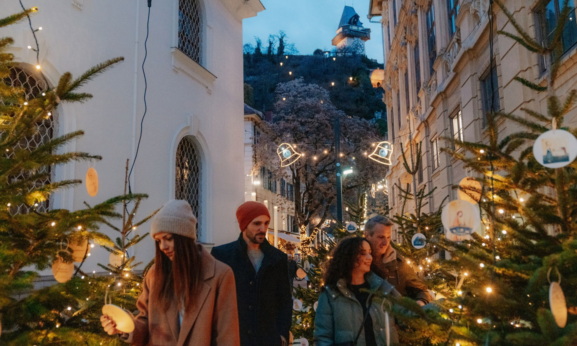 Vier Menschen spazieren zwischen Tannenbäumen durch die Grazer Innenstadt.