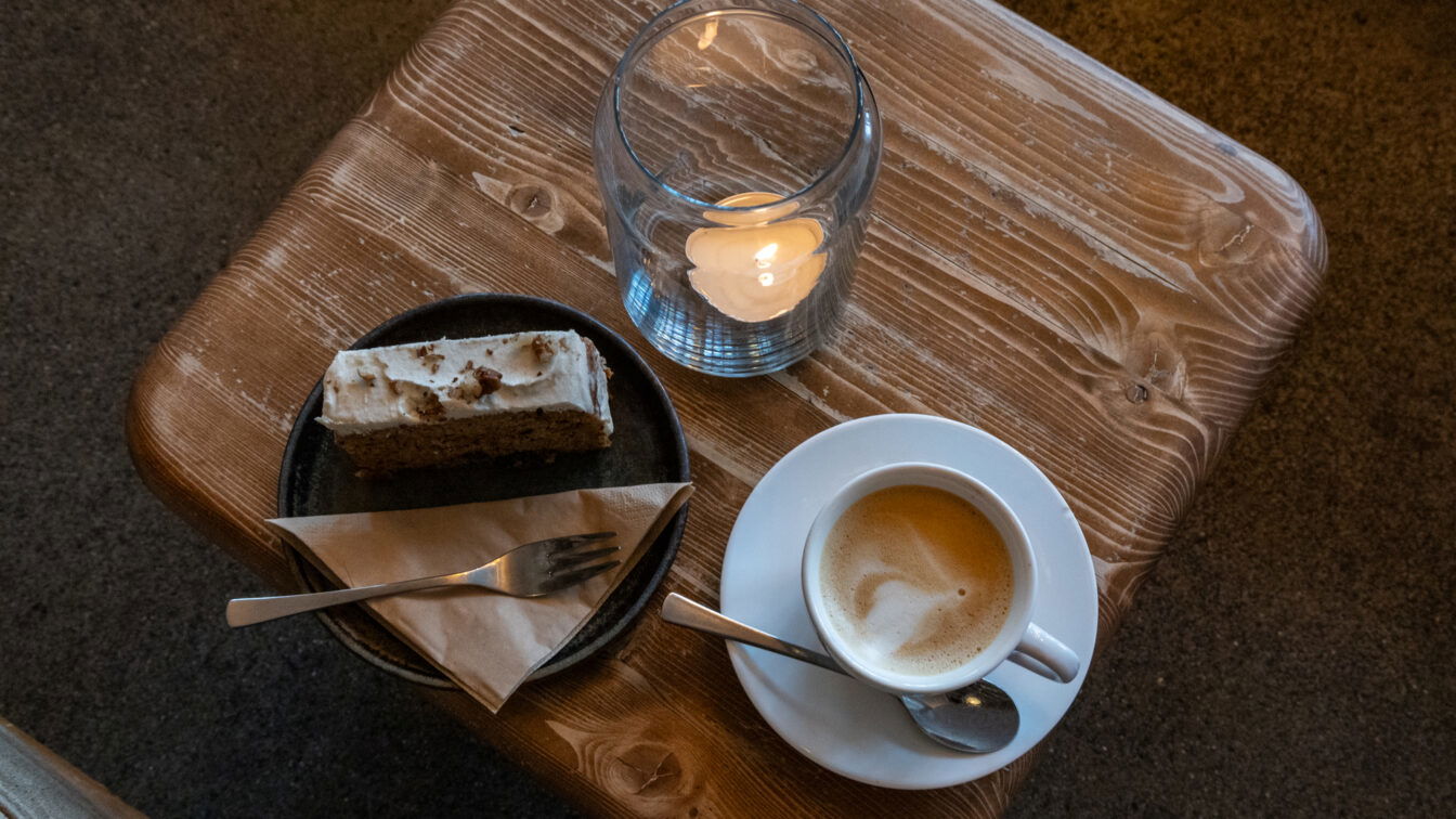 Eine Tasse Kaffee mit Milchschaum, ein Stück Kuchen auf einem Teller und eine brennende Kerze auf einem rustikalen Holztisch in gemütlicher Café-Atmosphäre in Berlin