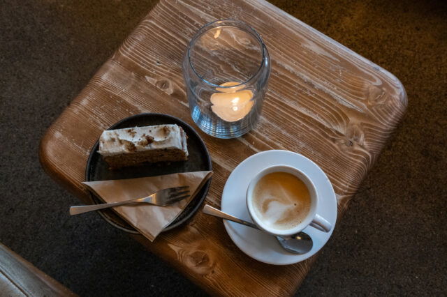 Eine Tasse Kaffee mit Milchschaum, ein Stück Kuchen auf einem Teller und eine brennende Kerze auf einem rustikalen Holztisch in gemütlicher Café-Atmosphäre in Berlin