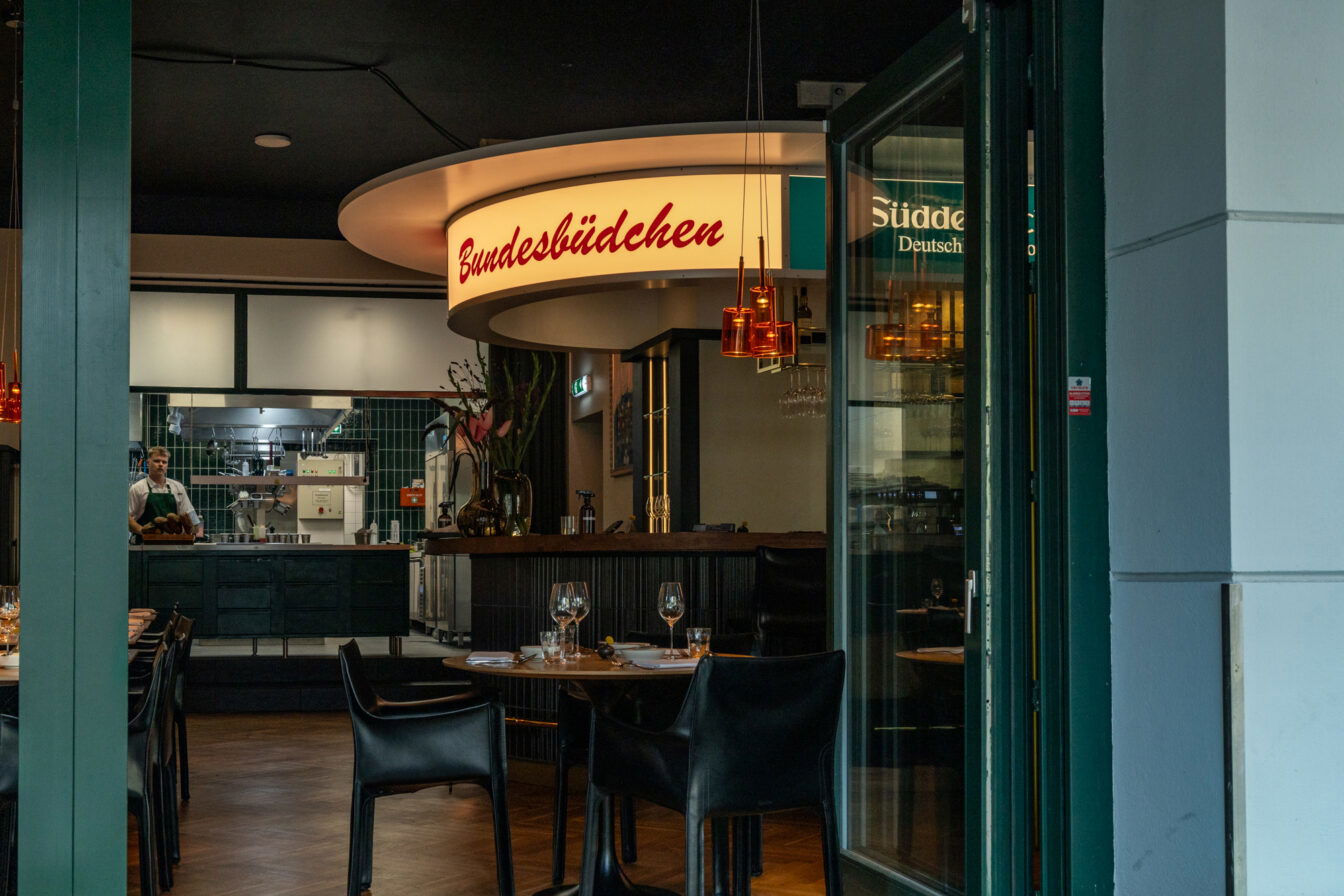 Blick ins Berliner Restaurant Bundesbüdchen mit stilvoll gedeckten Tischen, offener Küche und leuchtendem Retro-Schild, das Gemütlichkeit und moderne deutsche Gastronomie verbindet