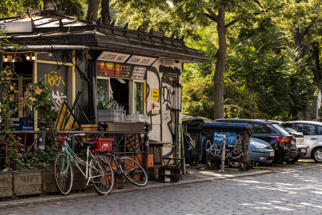 Straßenszene in Berlin-Kreuzberg mit einem kleinen, mit Graffiti verzierten Kiosk, Fahrrädern, Müllcontainern und parkenden Autos unter grünen Bäumen bei Sonnenschein.