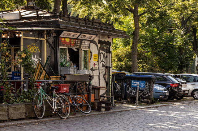 Straßenszene in Berlin-Kreuzberg mit einem kleinen, mit Graffiti verzierten Kiosk, Fahrrädern, Müllcontainern und parkenden Autos unter grünen Bäumen bei Sonnenschein.