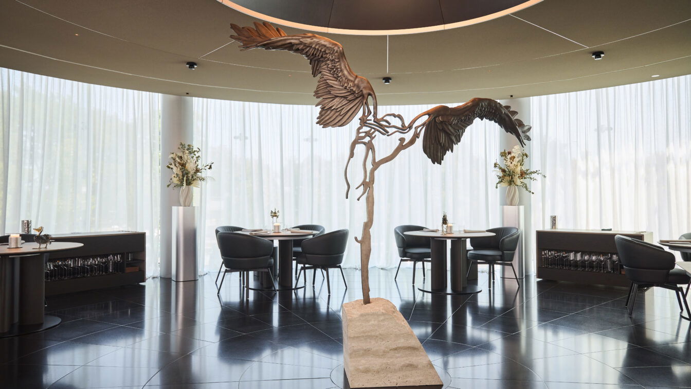 (c) Helge Kirchberger Photography | Red Bull Elegantes Fine-Dining-Restaurant mit moderner, runder Architektur und hohen, lichtdurchlässigen Vorhängen. Im Zentrum steht eine kunstvolle Skulptur mit ausgebreiteten Flügeln, die an den Ikarus-Mythos erinnert. Die Einrichtung ist stilvoll mit runden Tischen, dunklen Sesseln und dezenten Blumendekorationen.