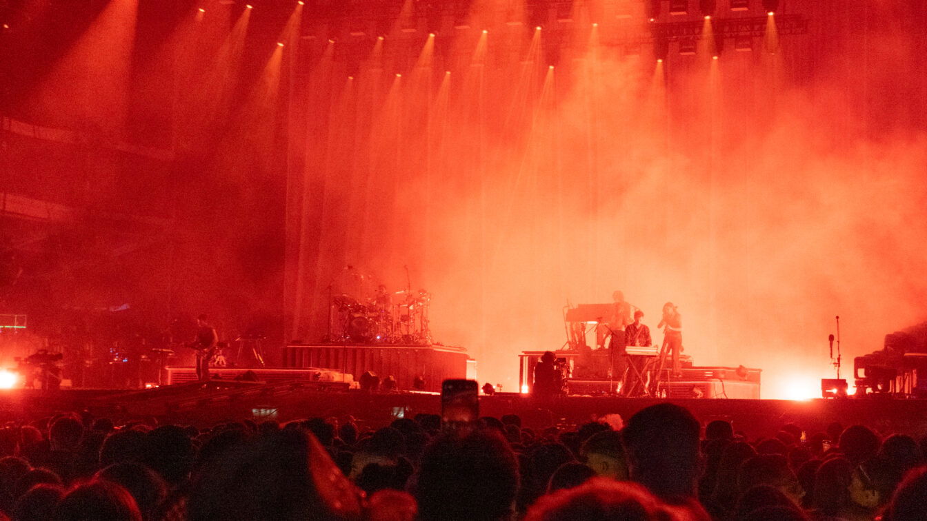 (c) Lilli Wermuth | 1000things Bühne bei einem Open-Air-Konzert in Berlin, in rotes Licht getaucht. Musikerinnen und Musiker spielen auf Instrumenten wie Schlagzeug, Keyboard und Gitarre, während dichtes Publikum davor steht. Nebel und Scheinwerfer verstärken die intensive Atmosphäre.