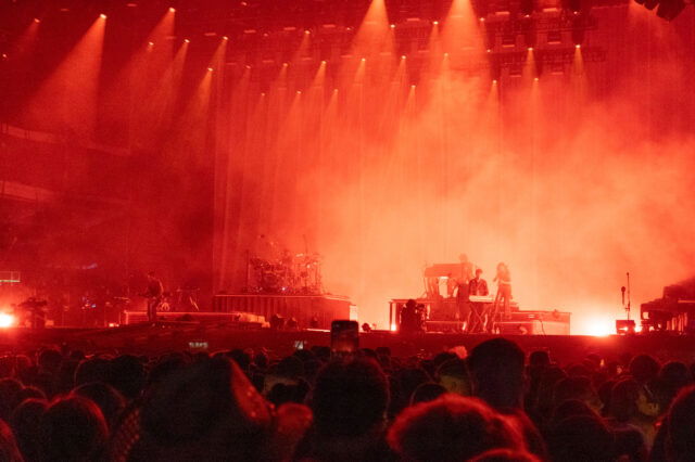 Bühne bei einem Open-Air-Konzert in Berlin, in rotes Licht getaucht. Musikerinnen und Musiker spielen auf Instrumenten wie Schlagzeug, Keyboard und Gitarre, während dichtes Publikum davor steht. Nebel und Scheinwerfer verstärken die intensive Atmosphäre.
