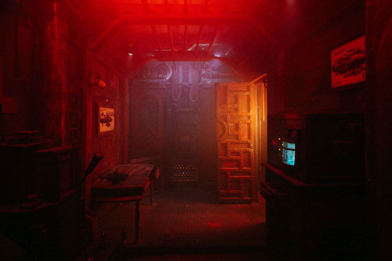 Eine Aktivität für Sonntage in Wien: Escape Game spielen. Ein schmaler, düster beleuchteter Raum mit industriellem und futuristischem Design, in rotem Licht getaucht. An den Wänden hängen technische Zeichnungen und Metallrohre verlaufen an der Decke. Rechts befindet sich ein Gerät mit leuchtendem blauen Bildschirm. Eine massive Tür aus Metall und Holz ist halb geöffnet und führt in einen weiteren, noch dunkleren Bereich.