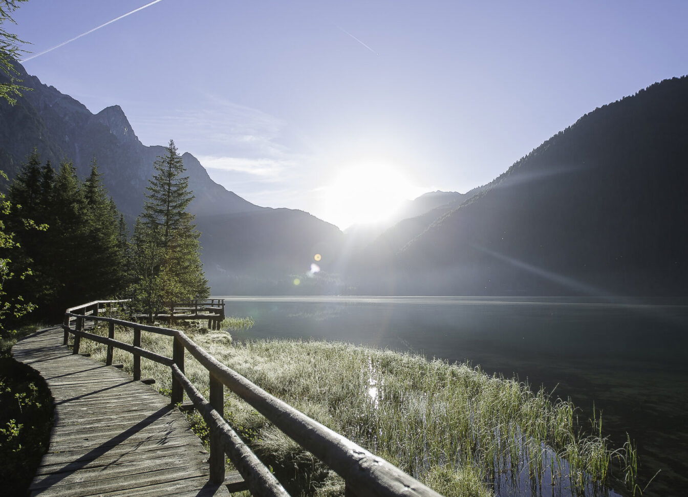 Holzsteg mit Geländer am Ufer des Antholzer Sees, gesäumt von Bäumen und Schilf, im Hintergrund Berge und Sonnenaufgang über den Gipfeln.