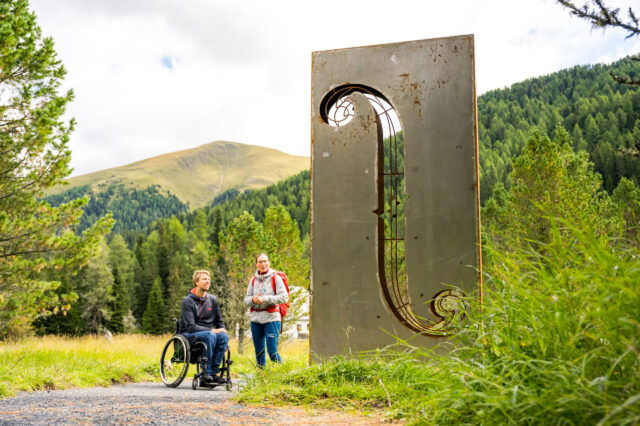 Eine Person im Rollstuhl und eine nicht beeinträchtigte Person vor einer Skulptur im Grünen.