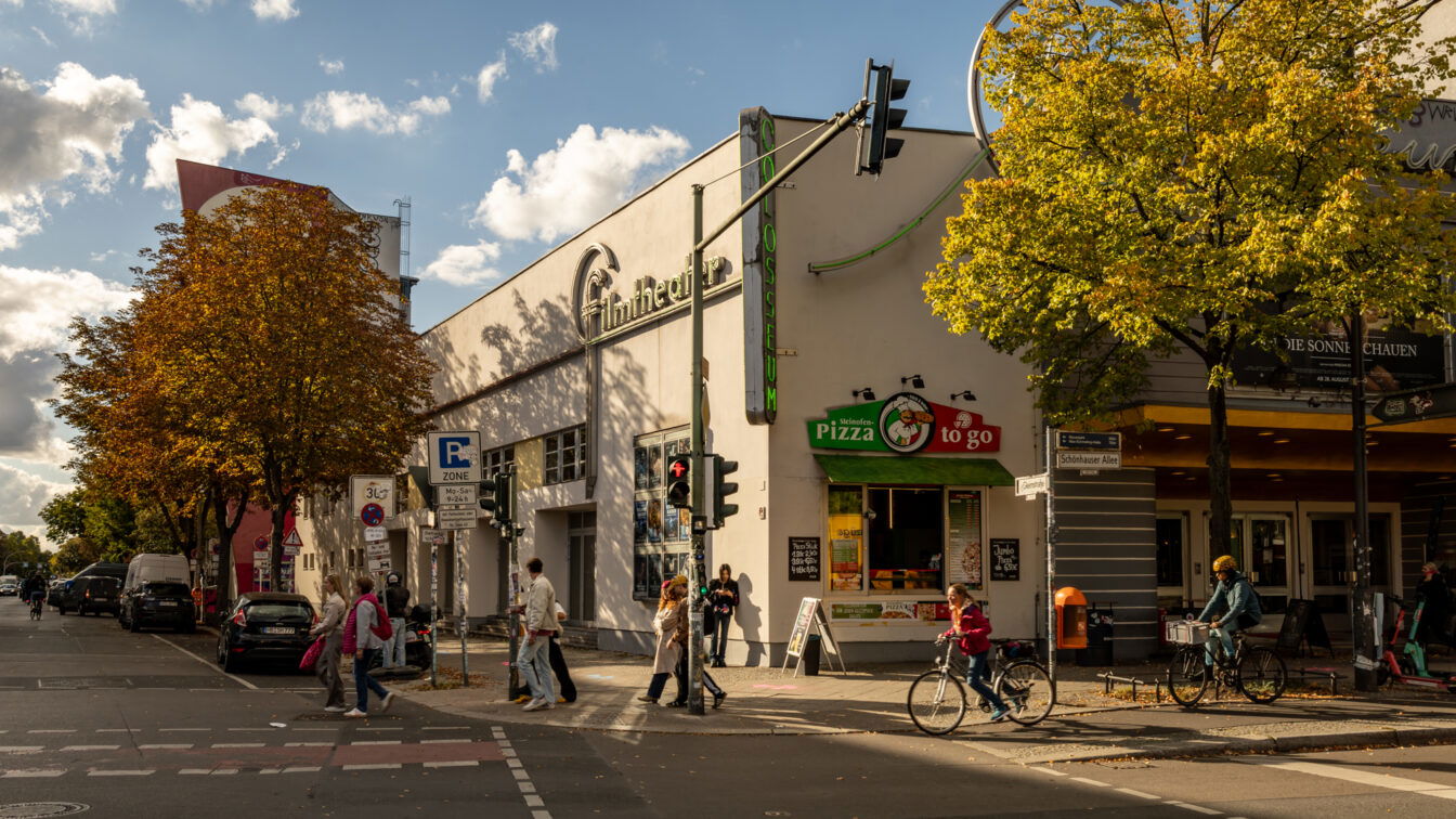 (c) Sonja Koller | 1000things Straßenszene in Berlin im Oktober: Fußgänger überqueren die Straße an einer Ampel, Radfahrer fahren vorbei. Herbstlich gefärbte Bäume mit gelben und orangefarbenen Blättern säumen den Straßenrand. Im Hintergrund das Gebäude des Filmtheaters Colosseum mit Leuchtreklame und einem Imbiss für Pizza to go.