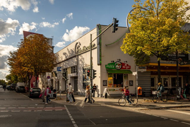 Straßenszene in Berlin im Oktober: Fußgänger überqueren die Straße an einer Ampel, Radfahrer fahren vorbei. Herbstlich gefärbte Bäume mit gelben und orangefarbenen Blättern säumen den Straßenrand. Im Hintergrund das Gebäude des Filmtheaters Colosseum mit Leuchtreklame und einem Imbiss für Pizza to go.