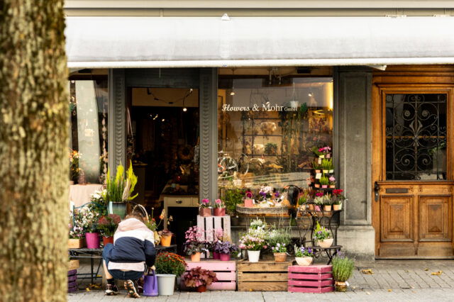Ein gemütlicher Blumenladen in Berlin mit dem Namen ‚Flowers & Mohr GmbH‘. Vor dem Laden stehen bunte Blumentöpfe und Pflanzen auf Holzkisten und kleinen Tischen arrangiert. Eine Person kniet draußen und kümmert sich um die Pflanzen. Der Eingang ist von einer weißen Markise und einer dekorativen Holztür flankiert.