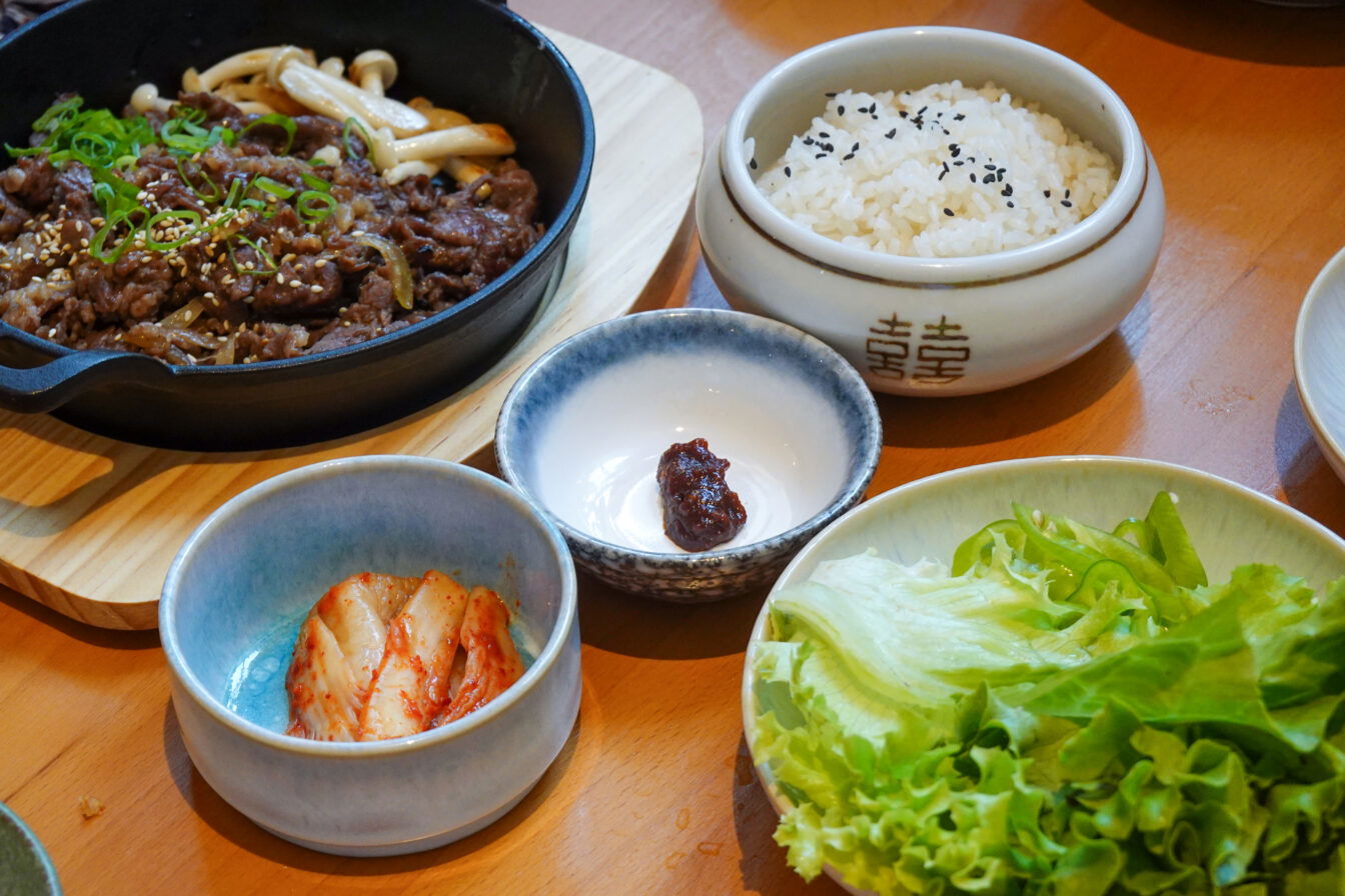 bulgogi bei dodo 62, mehrere schalen und schüsseln auf einem Tisch, reichlich befüllt mit Rindfleisch und Shimeji Pilzen, eine Schale mit Reis, eine mit Kimchi, eine mit einer dunklen Paste und eine mit Salat