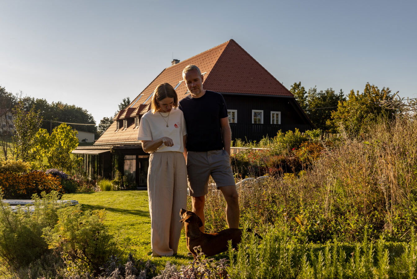 Ein Paar steht im blühenden Garten des Old School Guesthouse mit einem kleinen Hund, im Hintergrund das renovierte historische Gebäude mit rotem Ziegeldach.