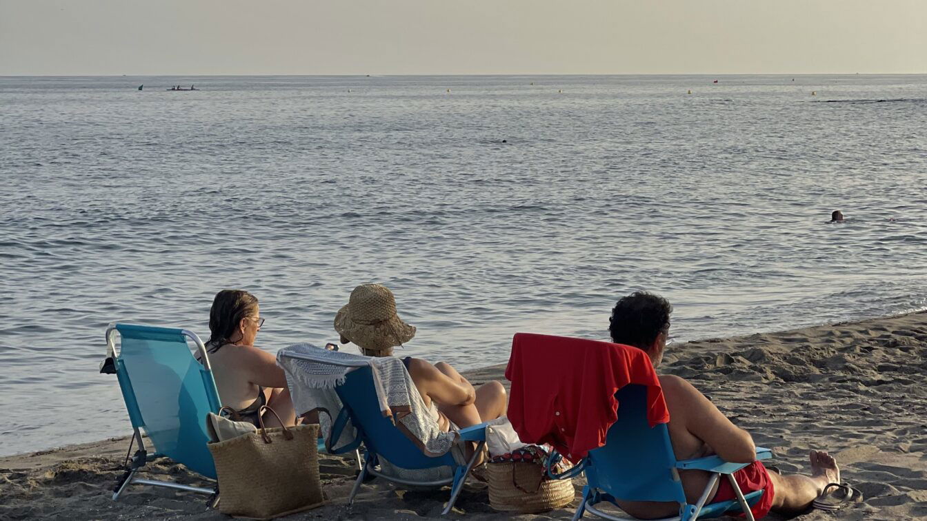 (c) Dupe | Maria Santos Drei Menschen sitzen auf Liegestühlen am Strand. Es ist Abendstimmung.