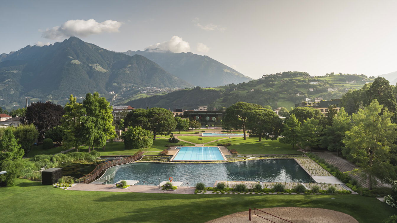 © KOTTERSTEGER Therme Meran in Südtirol mit Außenpools und weitläufiger Parkanlage, umgeben von grünen Hügeln und imposanten Alpen im Hintergrund – ideal für Wellness, Entspannung und Naturerlebnis.