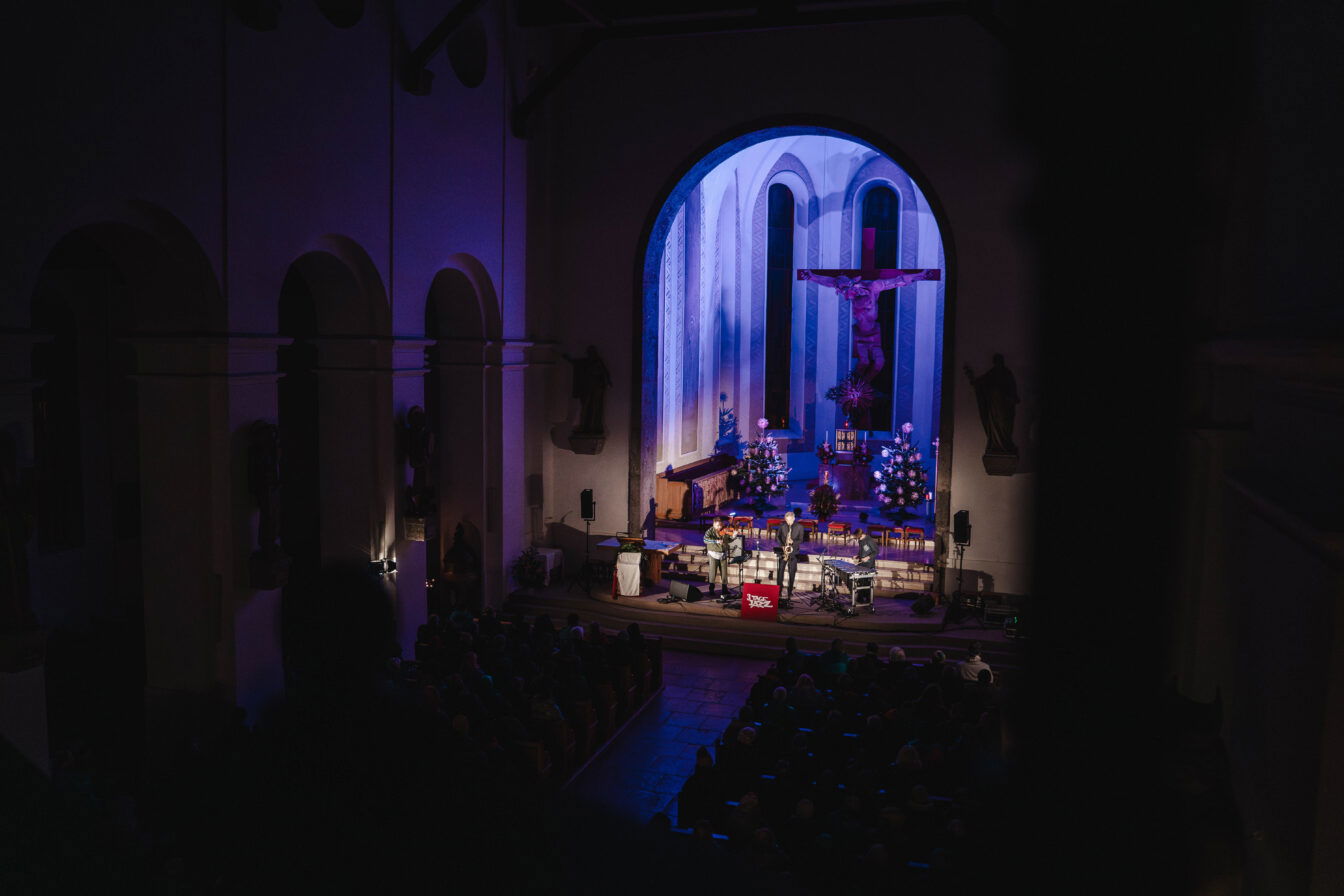 Konzert im Rahmen des Festivals 3 Tage Jazz in einer stimmungsvoll beleuchteten Kirche: Eine Jazzband spielt auf der Bühne vor dem Altarraum unter einem großen Kruzifix, während das Publikum in den Kirchenbänken sitzt und zuhört.