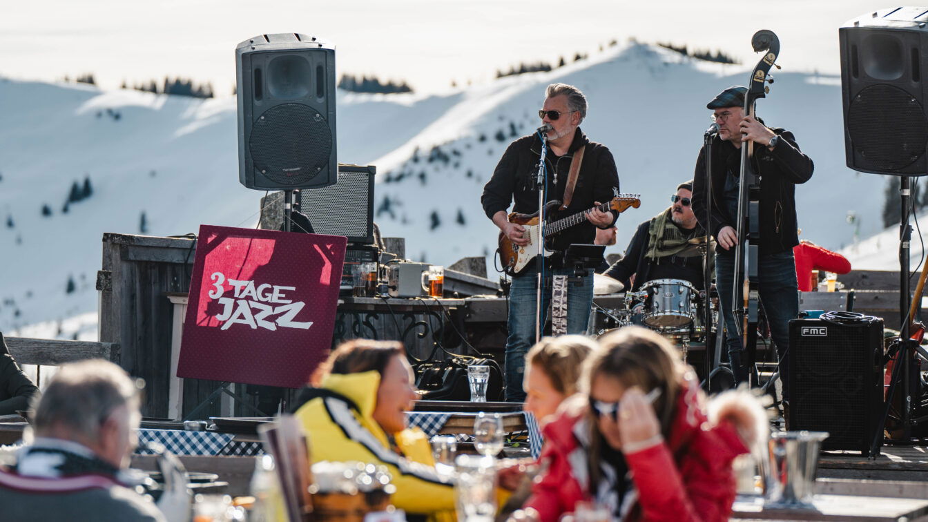 (c) Michael Geißler Open-Air-Jazzkonzert im Rahmen des Festivals 3 Tage Jazz in Saalfelden: Eine Band spielt mit Gitarre, Kontrabass und Schlagzeug vor verschneiten Berggipfeln, während Besucher in Winterkleidung an Holztischen sitzen und Getränke genießen.