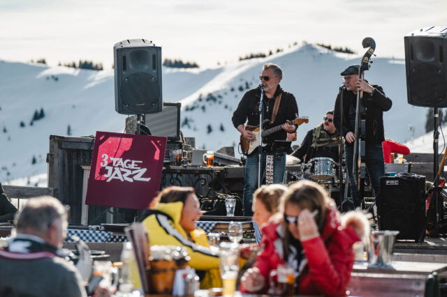 Open-Air-Jazzkonzert im Rahmen des Festivals 3 Tage Jazz in Saalfelden: Eine Band spielt mit Gitarre, Kontrabass und Schlagzeug vor verschneiten Berggipfeln, während Besucher in Winterkleidung an Holztischen sitzen und Getränke genießen.