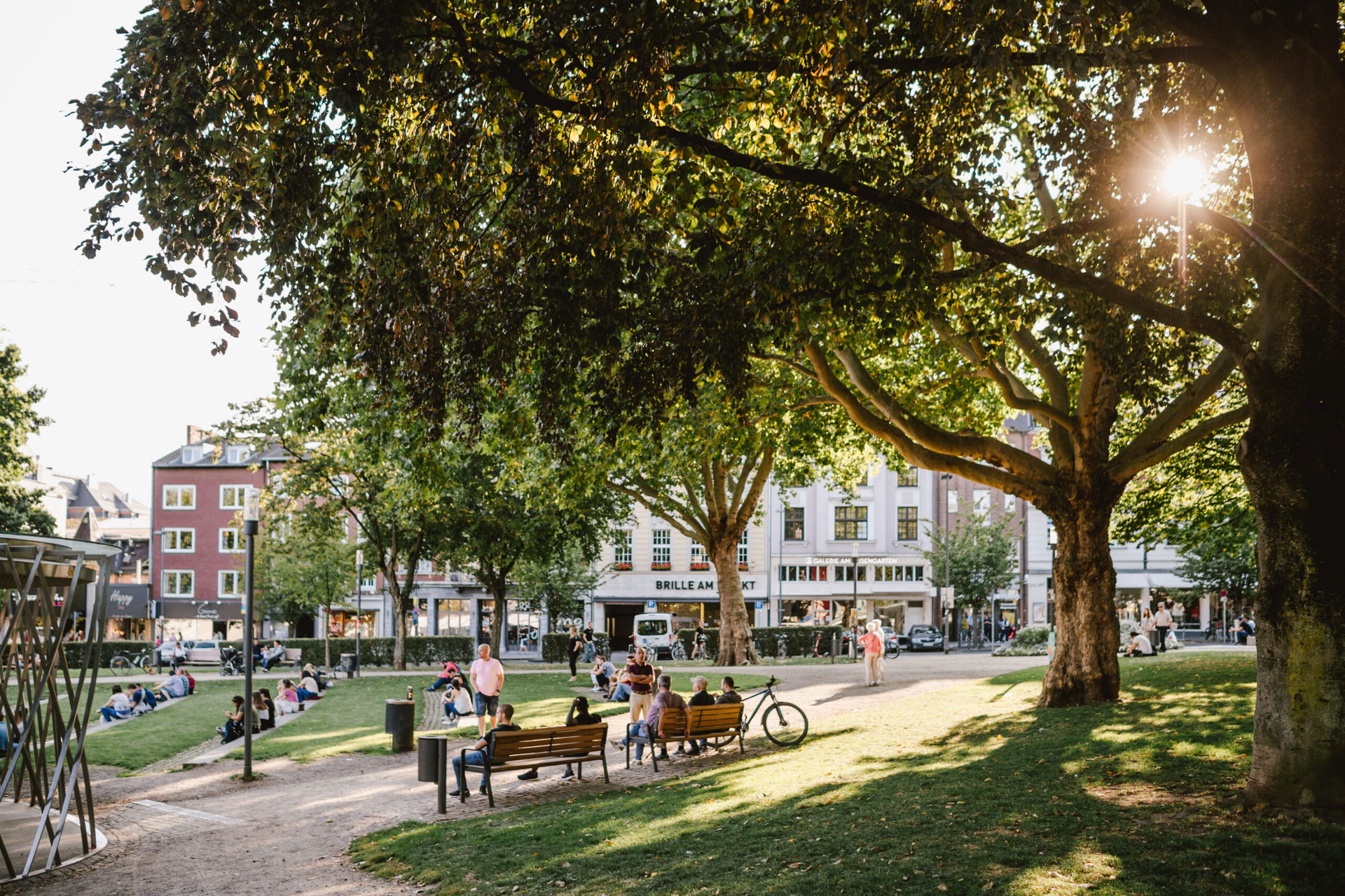Menschen genießen den sonnigen Nachmittag im Elisengarten in Aachen, sitzen auf Bänken und im Gras unter großen, schattenspendenden Bäumen mit Blick auf umliegende Altstadthäuser.