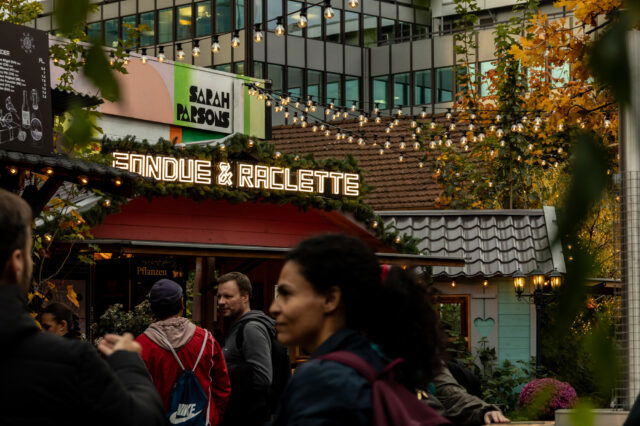 (c) Sonja Koller | 1000things Belebte Adventsszene in Zürich mit einem festlich geschmückten Stand, auf dem in leuchtenden Buchstaben ‚Fondue & Raclette‘ steht. Menschen schlendern davor entlang, während über ihnen Lichterketten hängen. Im Hintergrund sind herbstlich gefärbte Bäume und moderne Gebäude zu sehen.