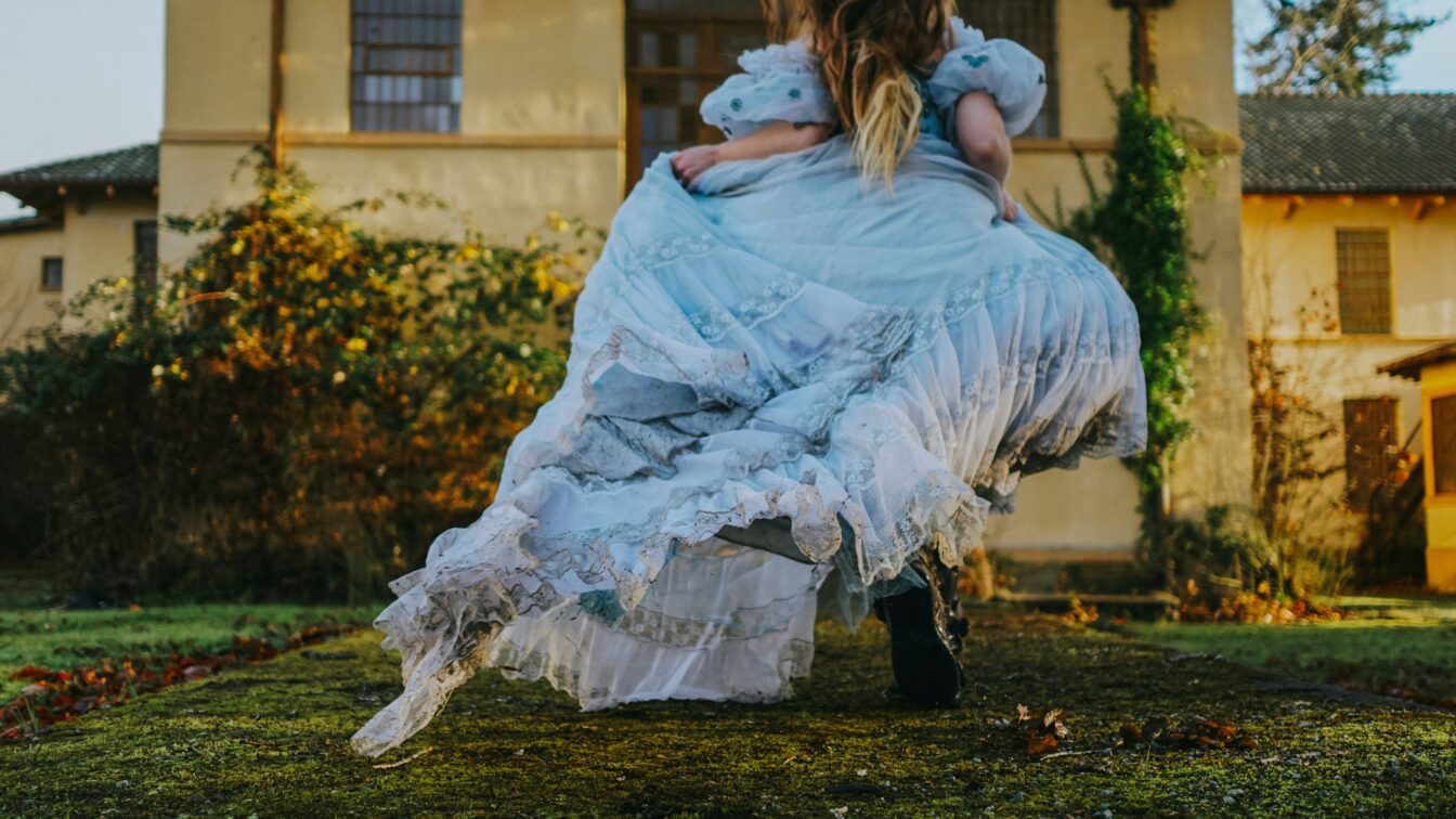 (c) Elle Cartier | unsplash Eine Person in einem langen, hellblauen, verspielten Kleid läuft oder tanzt über einen moosbewachsenen Weg vor einem alten Gebäude. Der Stil erinnert an ein Märchen oder eine Szene aus Alice im Wunderland.