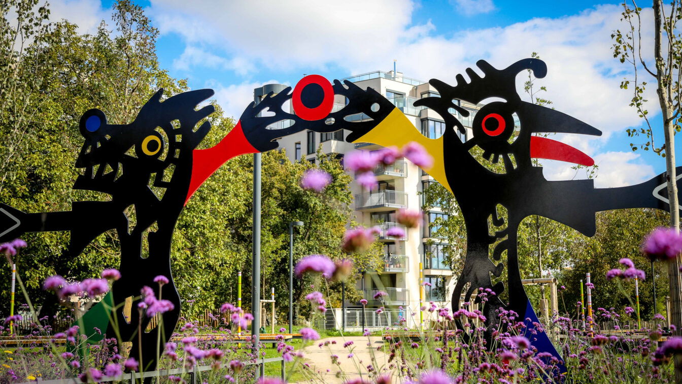 Farbenfrohe Skulpturen von André Heller im Kunstpark in Wien, eingerahmt von blühenden Blumen und moderner Architektur im Hintergrund.