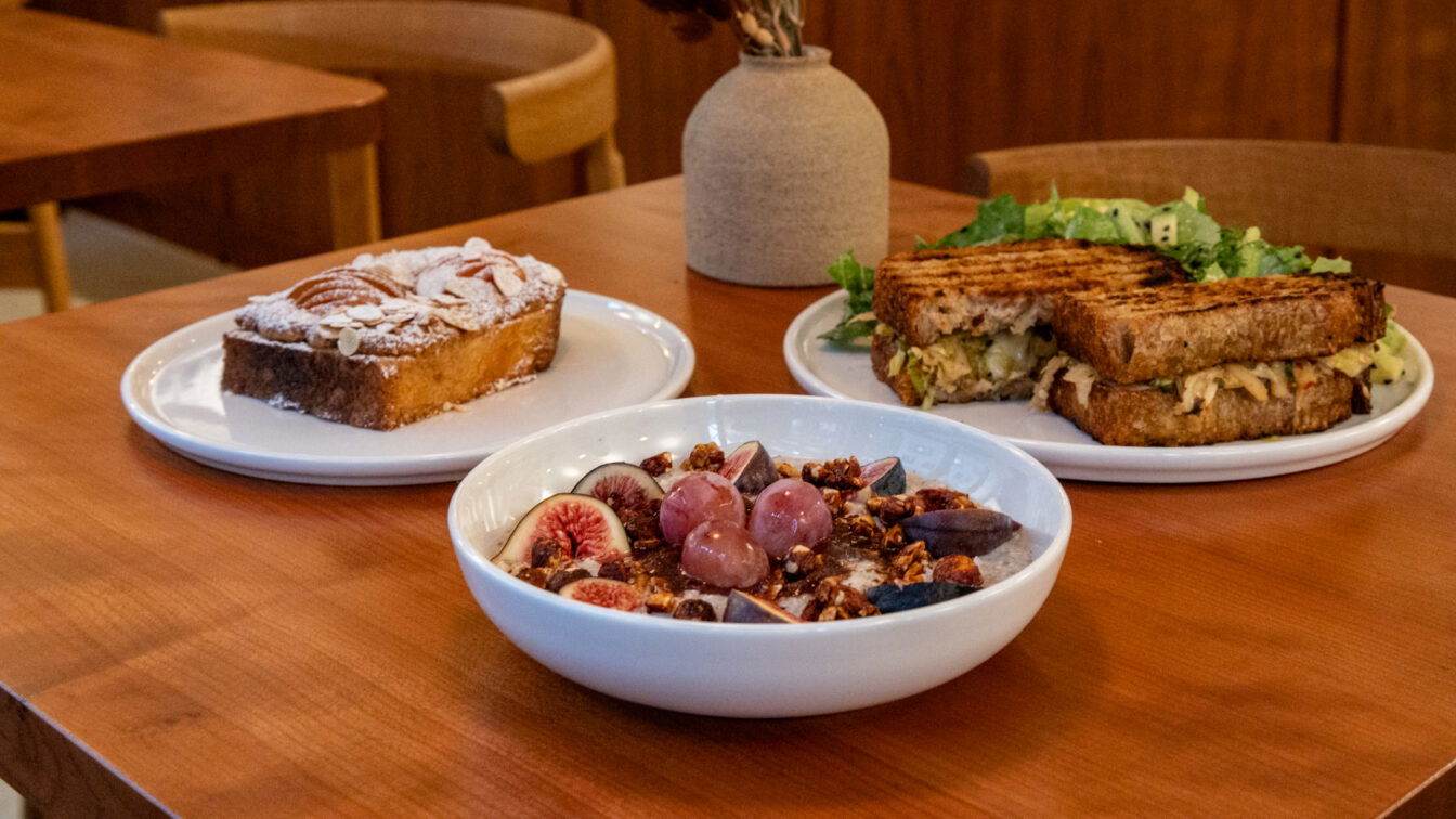 (c) Sonja Koller | 1000things Ein gemütlich gedeckter Holztisch in einem Café in Zürich mit drei Brunchgerichten: ein Teller mit einem Stück Mandelkuchen, ein Teller mit einem gegrillten Sandwich und Salat, sowie eine Schüssel mit Porridge, frischen Feigen, Trauben und Granola. Im Hintergrund stehen Holzstühle und eine Vase mit Trockenblumen.