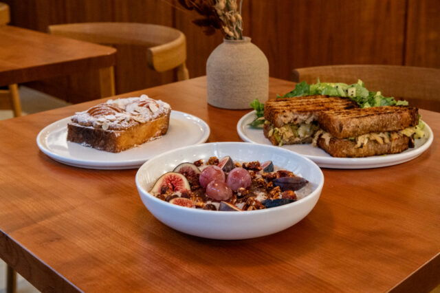 Ein gemütlich gedeckter Holztisch in einem Café in Zürich mit drei Brunchgerichten: ein Teller mit einem Stück Mandelkuchen, ein Teller mit einem gegrillten Sandwich und Salat, sowie eine Schüssel mit Porridge, frischen Feigen, Trauben und Granola. Im Hintergrund stehen Holzstühle und eine Vase mit Trockenblumen.