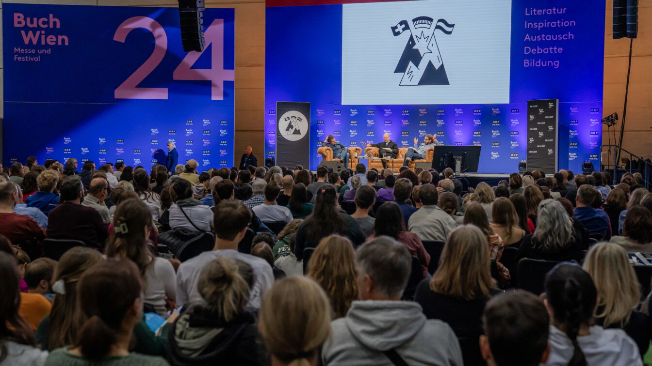 Publikum bei einer Podiumsdiskussion auf der Buch Wien 2024. Auf der Bühne sitzen mehrere Personen in Sesseln, hinter ihnen eine große Leinwand mit Logo und Schriftzug. Links und rechts sind große blaue Banner mit der Aufschrift ‚Buch Wien – Messe und Festival‘ sowie ‚Literatur, Inspiration, Austausch, Debatte, Bildung‘