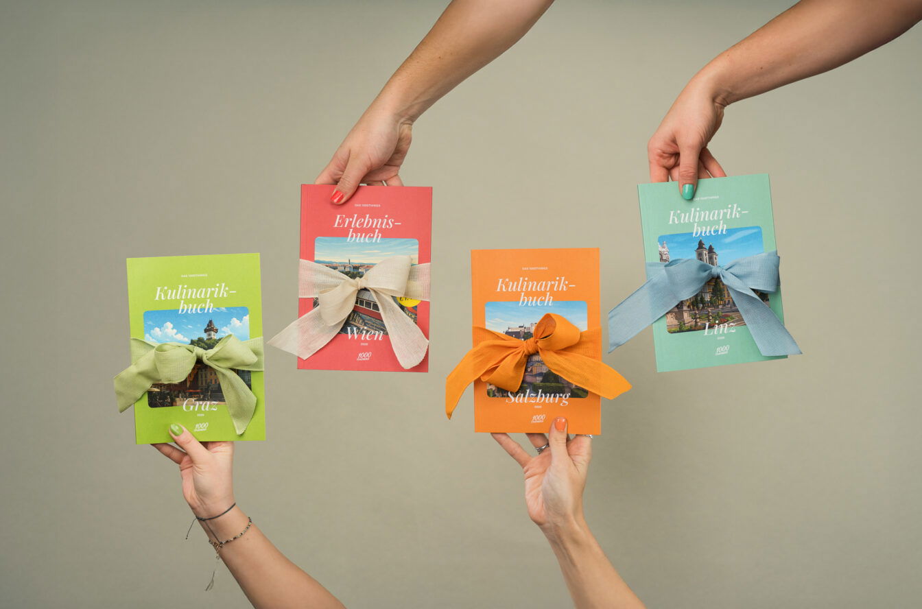 Vier Hände halten die 1000things Bücher – Erlebnisbuch Wien und Kulinarikbücher Graz, Salzburg und Linz – jeweils mit farbigen Geschenk-Schleifen.