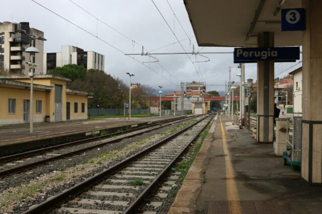 Bahngleise und Bahnsteig am Bahnhof Perugia in Italien, mit Gebäuden im Hintergrund und bewölktem Himmel.