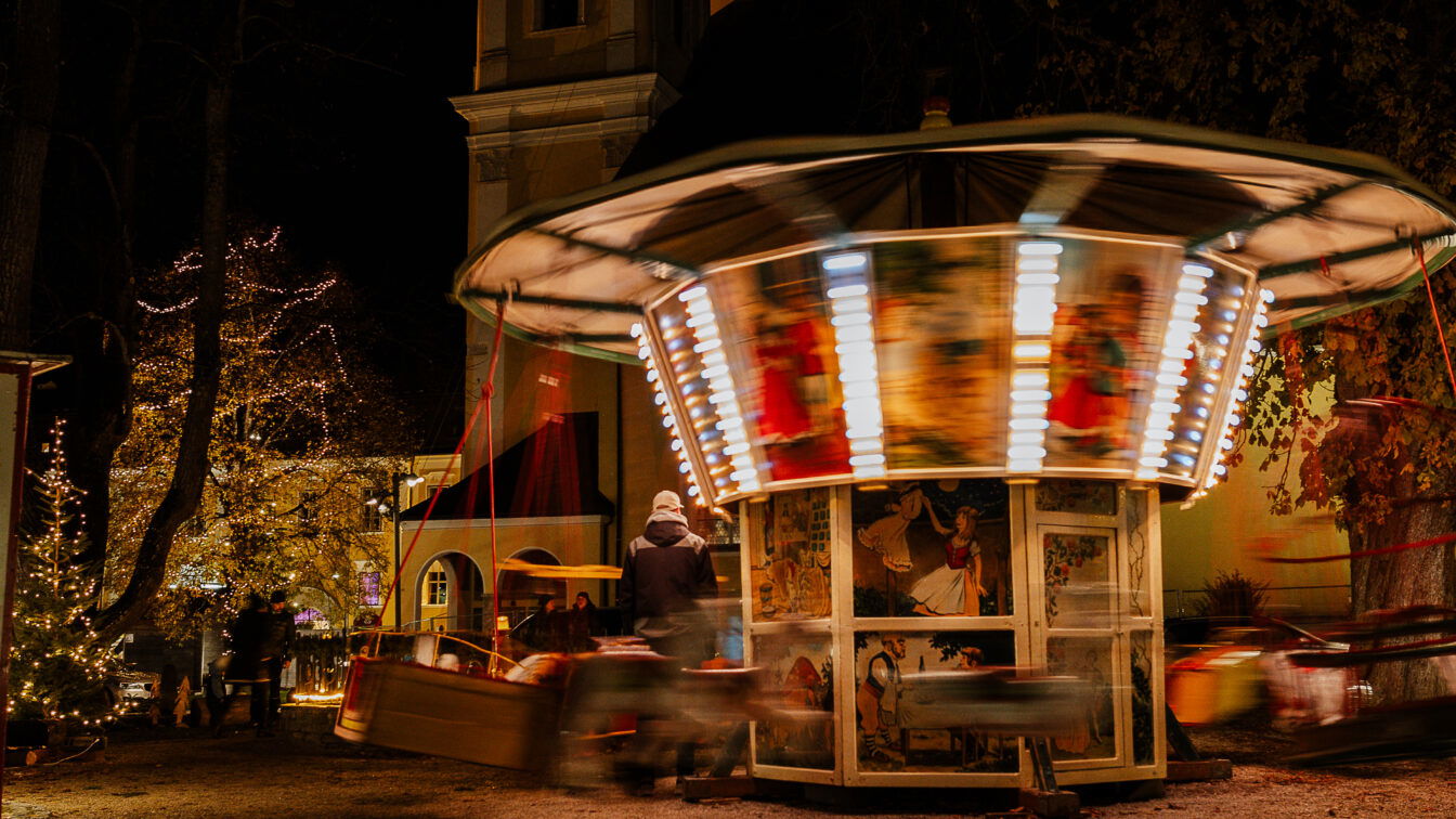 (c) Tourismusverband Mondsee-Irrsee | Kathrin Haas Nostalgisches Kinderkarussell mit bunten Lichtern dreht sich bei Nacht auf dem Adventmarkt in Mondsee, umgeben von festlich beleuchteten Bäumen und weihnachtlicher Atmosphäre.