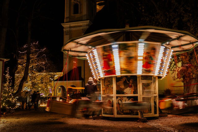 Nostalgisches Kinderkarussell mit bunten Lichtern dreht sich bei Nacht auf dem Adventmarkt in Mondsee, umgeben von festlich beleuchteten Bäumen und weihnachtlicher Atmosphäre.