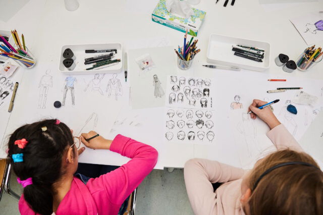 Zwei Kinder sitzen an einem Tisch und zeichnen Figuren und Porträts auf Papier. Auf dem Tisch liegen viele Bunt- und Bleistifte, Fineliner, Radiergummis und Skizzenblätter mit verschiedenen Körperhaltungen und Frisuren. Die Szene wirkt kreativ und konzentriert – ein Moment aus einem Kinderkunstfest oder Zeichenworkshop.