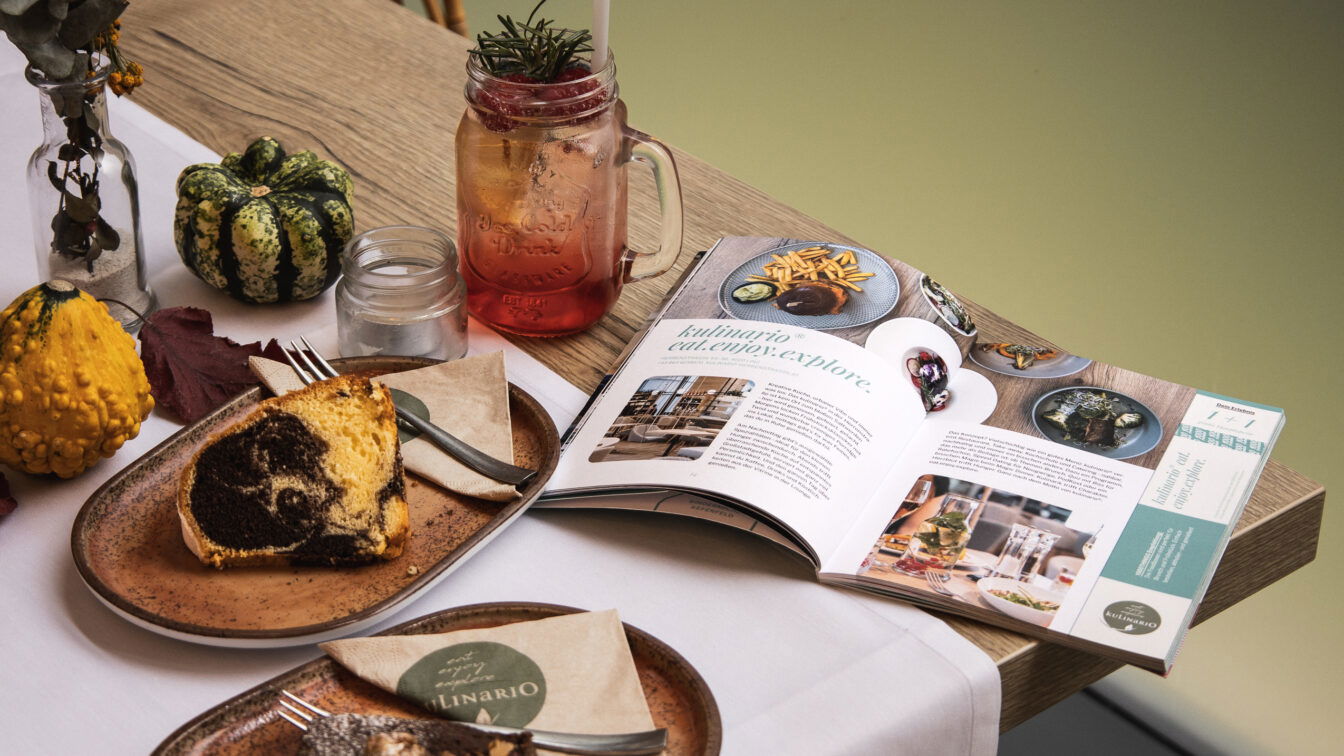 Auf einem gedeckten Tisch mit Kuchen, Getränk und herbstlicher Deko liegt ein geöffnetes 1000things Kulinarikbuch Linz.