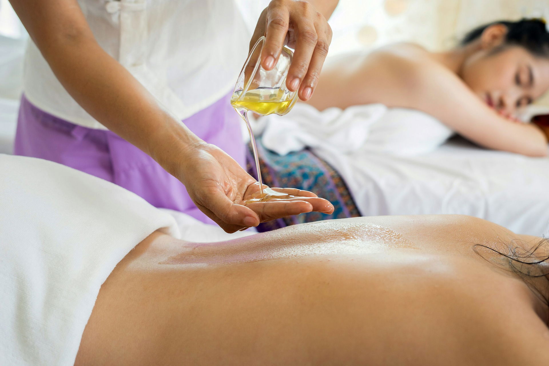 Wellness-Massage in Berlin: Therapeutin gießt warmes Massageöl auf den Rücken einer entspannten Kundin in einem hellen Spa