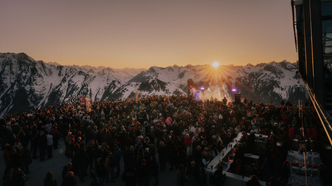 Eine große Menschenmenge feiert bei Sonnenuntergang auf einer Bergplattform umgeben von schneebedeckten Alpen. Im Vordergrund steht eine Bar, im Hintergrund eine Bühne mit Lichtshow. Das Bild zeigt die festliche Atmosphäre eines Ski-Openings 2025 in einem österreichischen Skigebiet