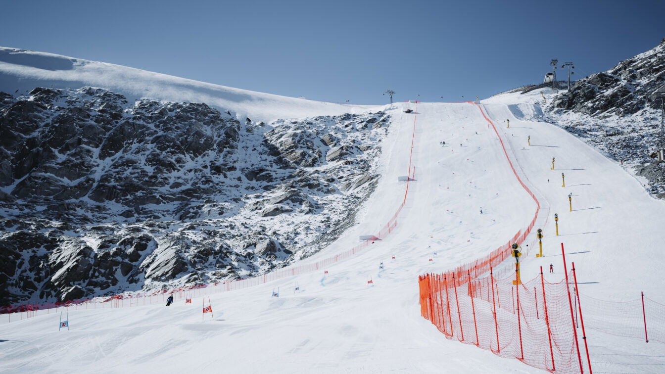 (c) Ötztal Tourismus | Tobias Siegele Ein schneebedeckter Hang im Skigebiet Sölden mit roter Sicherheitsabsperrung und Slalomtoren für ein Weltcuprennen. Mehrere Skifahrer sind auf der präparierten Piste unterwegs, umgeben von felsigen Berghängen unter blauem Himmel.