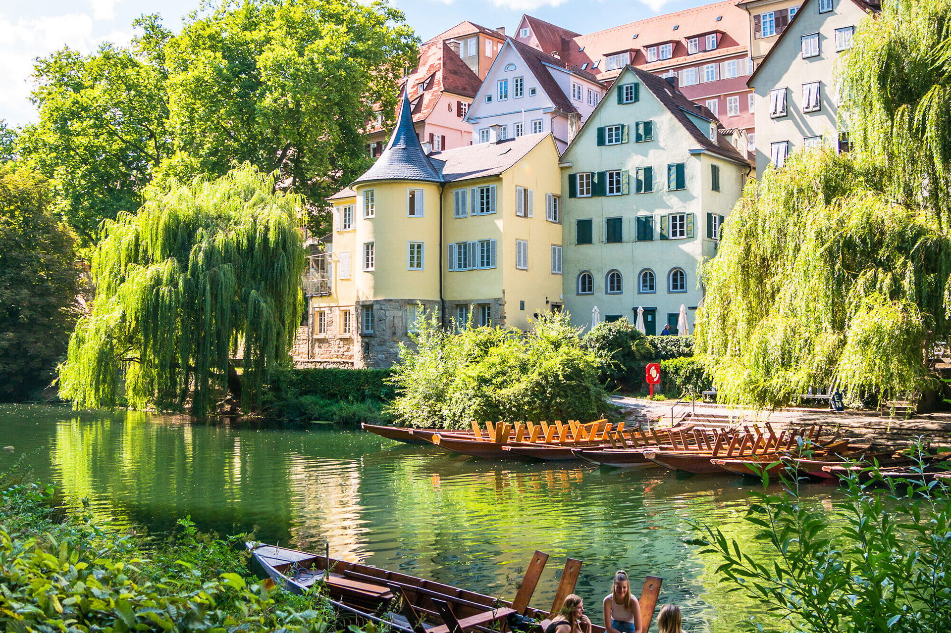 Blick auf den Hölderlinturm und bunte Altstadthäuser am Neckarufer in Tübingen, umgeben von üppigem Grün; im Vordergrund sitzen Menschen in einem Stocherkahn auf dem Wasser bei sonnigem Wetter.