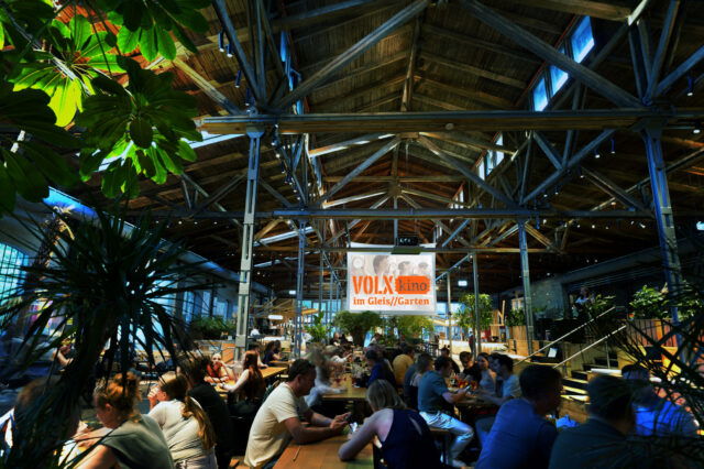 Innenaufnahme des VOLXkino im Gleis/Garten 2025: Menschen sitzen an langen Holztischen in einer großen, offenen Halle mit Holzdachkonstruktion, viel Grünpflanzen und gemütlicher Atmosphäre.