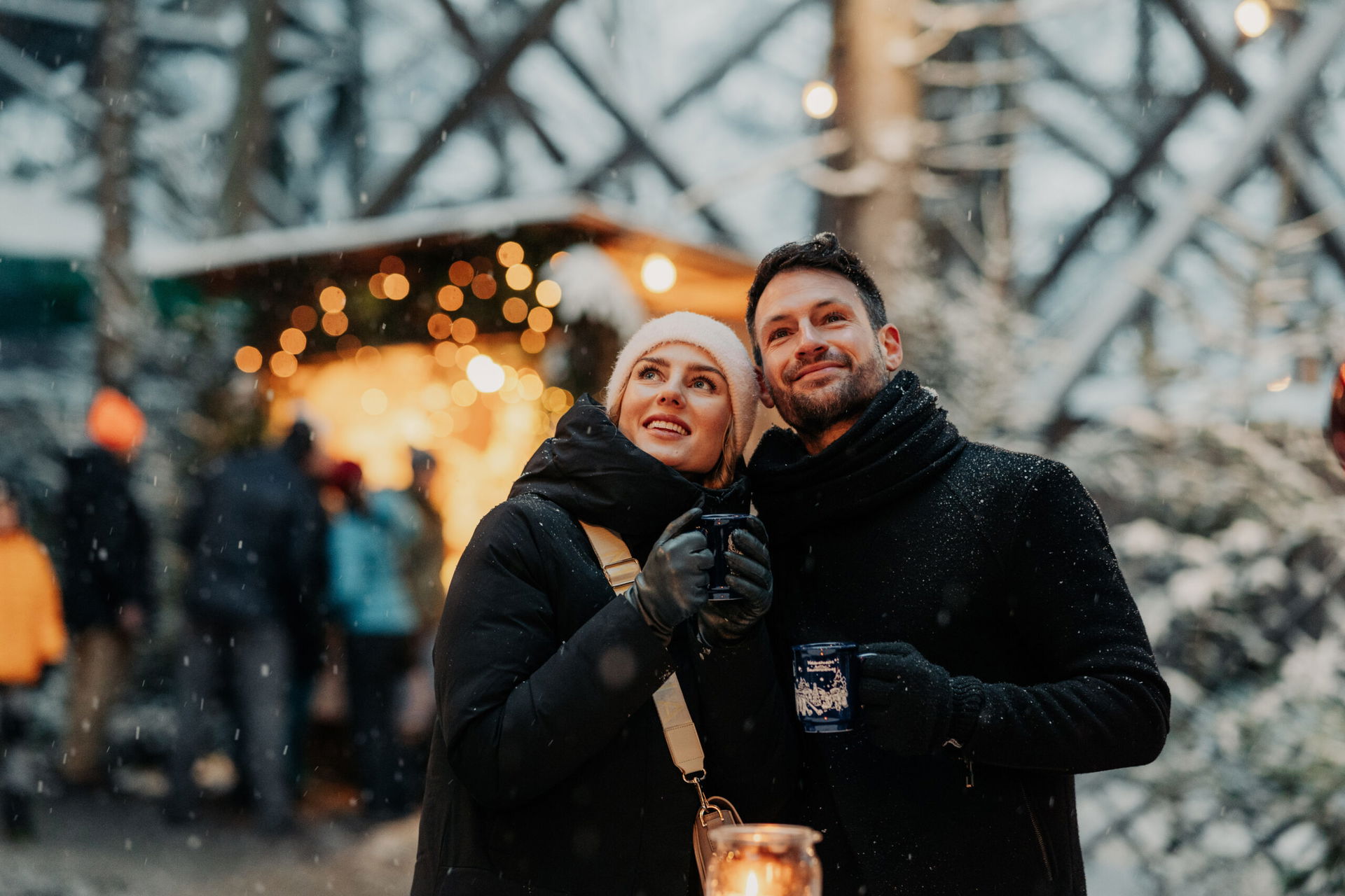 Ein verliebtes Paar genießt gemeinsam seinen Punsch oder Glühwein und lässt seinen Blick verträumt über die atemberaubende Umgebung der Waldweihnacht Kopfing schweifen, während es sich von der festlichen Stimmung und dem Zauber des Augenblicks verzaubern lässt. (c) CM Visuals