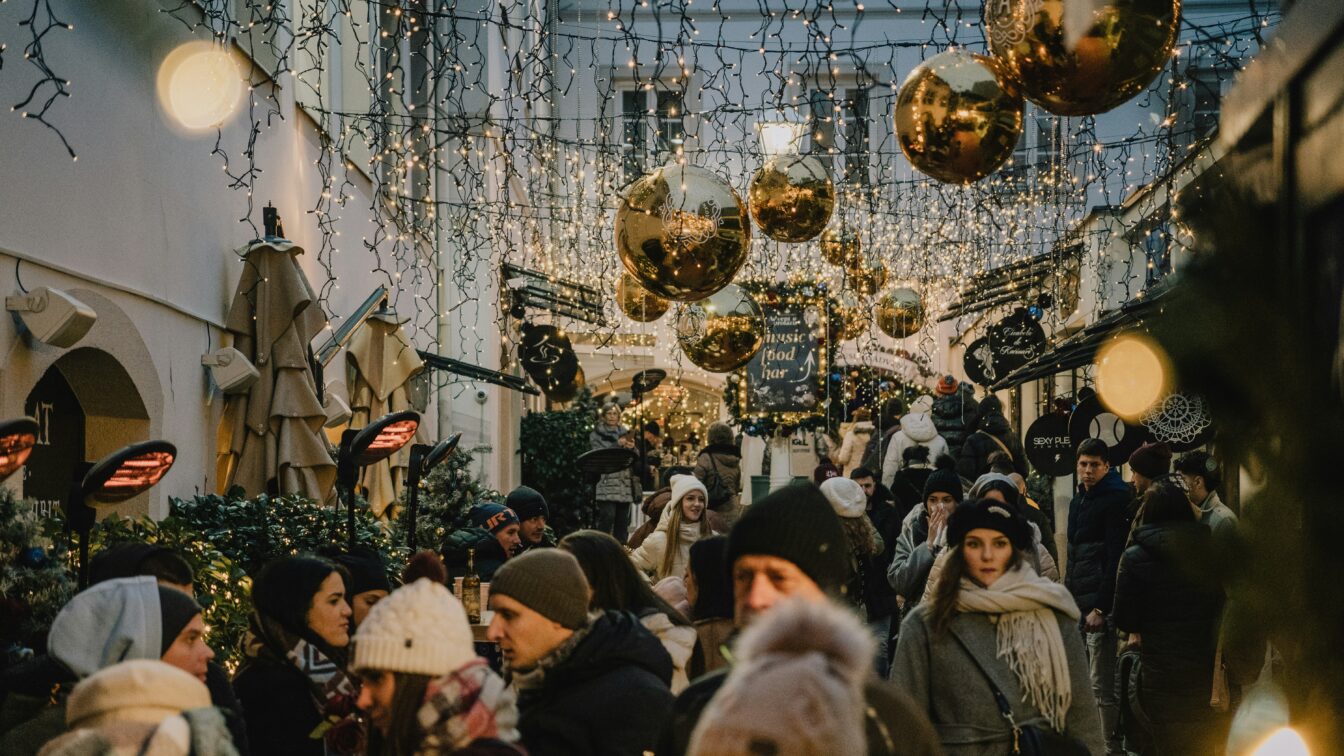 (c) Emmanuel Cassar | unsplash Belebter Weihnachtsmarkt in einer festlich geschmückten Gasse in Italien mit Lichterketten, goldenen Kugeln und Menschen in Winterkleidung.