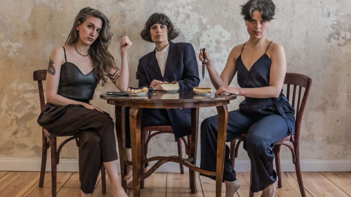 (c) Cristina Marx | Photomusix Drei Personen sitzen barfuß um einen kleinen Holztisch in einem schlichten Raum mit abgenutzter Wand. Sie blicken ernst in die Kamera. Auf dem Tisch liegen Teller mit Kuchenstücken und ein Messer, das eine der Personen in der Hand hält. Die Szene wirkt inszeniert und theatralisch, mit einem Hauch von Spannung