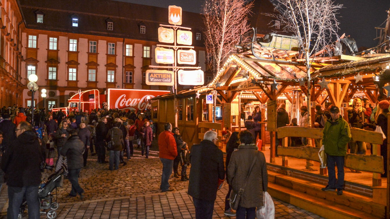 (c) Bayreuther Winterdorf Menschen genießen einen winterlichen Abend im Bayreuther Winterdorf, mit festlich beleuchteten Holzhütten, geschmückten Bäumen und einem leuchtenden Coca-Cola-Weihnachtstruck im Hintergrund.