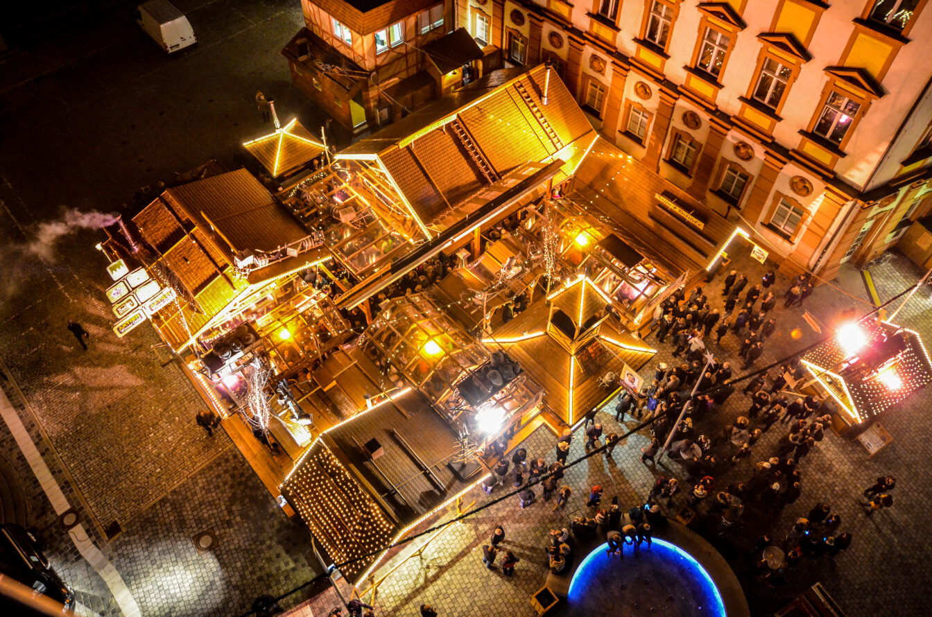 Luftaufnahme des Bayreuther Winterdorfs bei Nacht: zahlreiche beleuchtete Holzstände, festliche Lichterketten und Besucher, die durch die gemütlich erleuchteten Gassen schlendern.